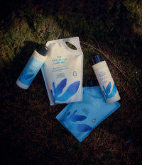 The Unscented Company nourishing shampoo and conditioner bottles with matching refill pouches, made with mango butter and argan oil, arranged on a mossy forest floor.