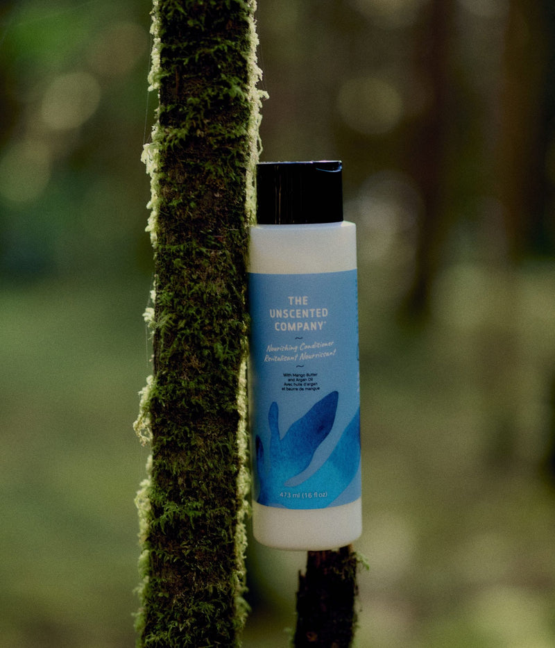 A bottle of The Unscented Company nourishing conditioner with mango butter and argan oil rests against a moss-covered tree in a lush green forest.