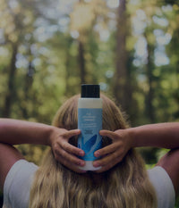 A blonde woman seen from behind holds a bottle of The Unscented Company moisturizing conditioner over her face while standing in a sunlit green forest.