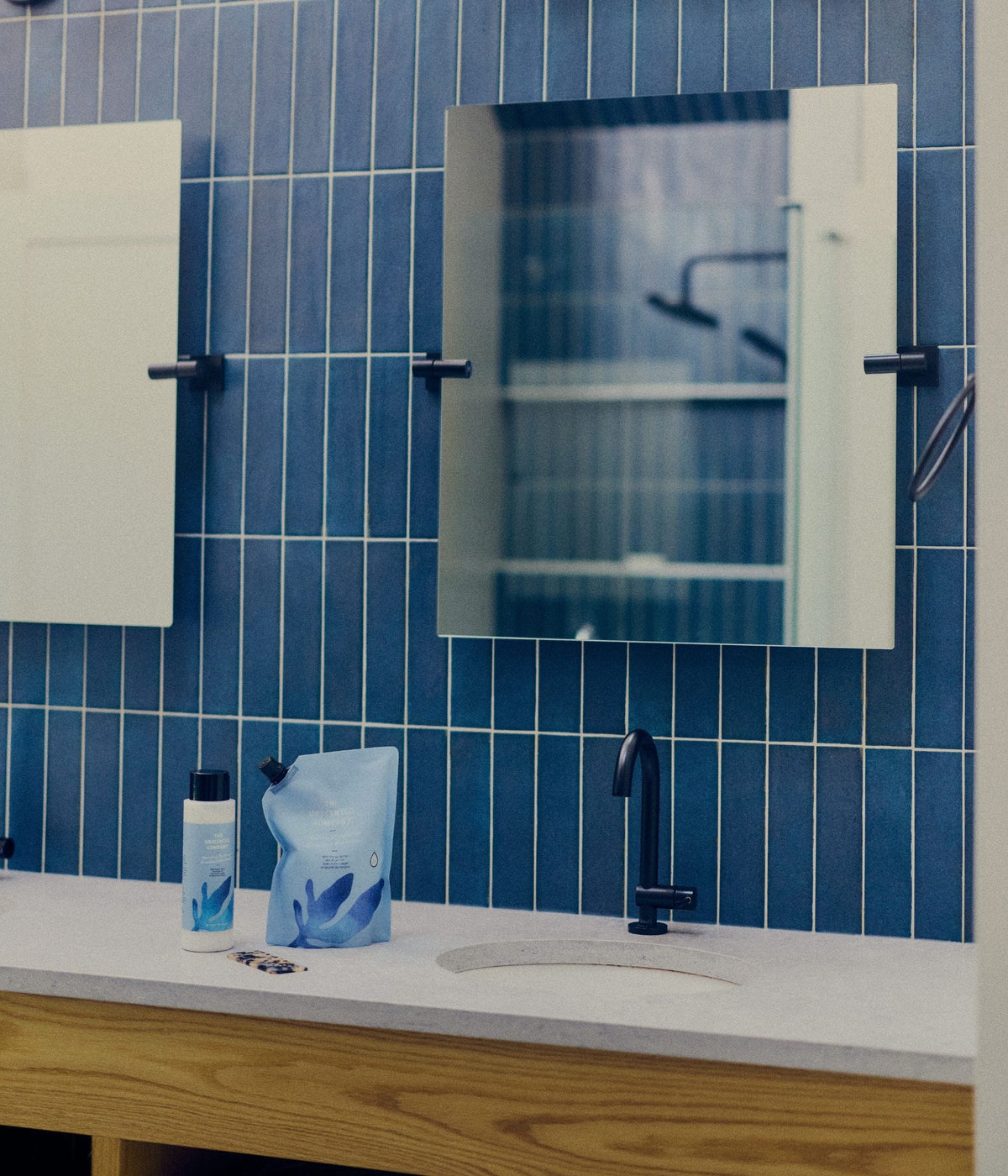 A bottle and refill pouch of The Unscented Company hair conditioner sit beside a white bathroom sink with a black faucet, blue tiled wall, and mirror above.