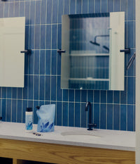 A bottle and refill pouch of The Unscented Company hair conditioner sit beside a white bathroom sink with a black faucet, blue tiled wall, and mirror above.