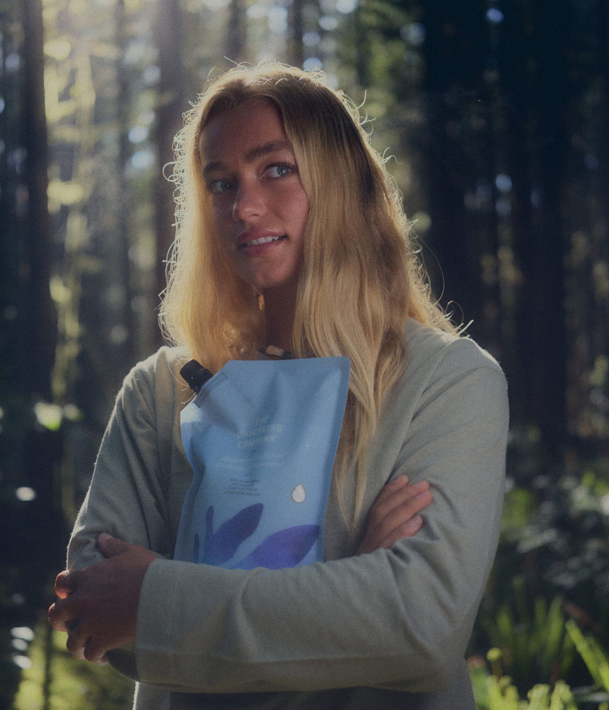 A blonde woman in a light green long-sleeve top stands in a sunlit forest, holding a blue The Unscented Company hair care refill pouch against her chest.