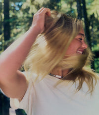 A smiling blonde woman in a white T-shirt runs her hand through her hair outdoors in a sunlit green forest, with her hair moving around her face.