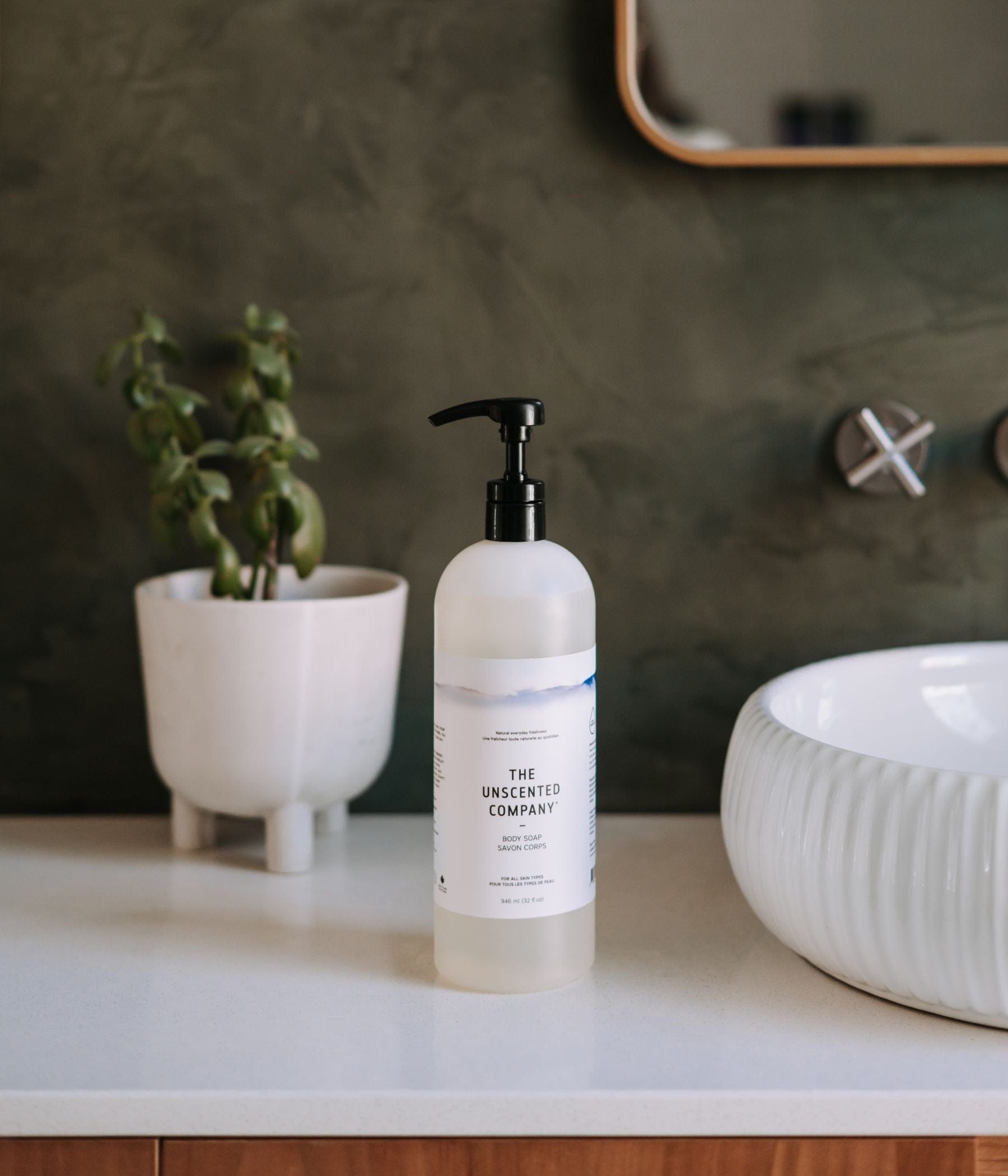 The Body Soap - 1L placed a bathroom counter, on the left side of a sink, dark green background.