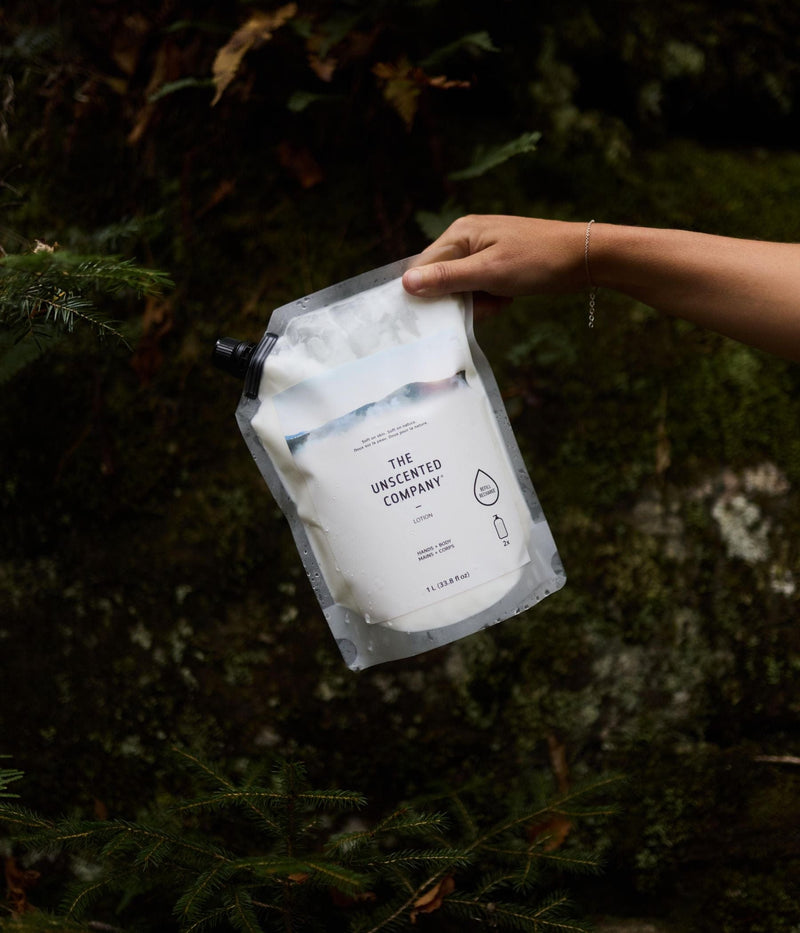 A hand holding a 1-liter refill pouch of The Unscented Company lotion in front of a mossy rock wall. The pouch label reads “Lotion – Hands + Body.”