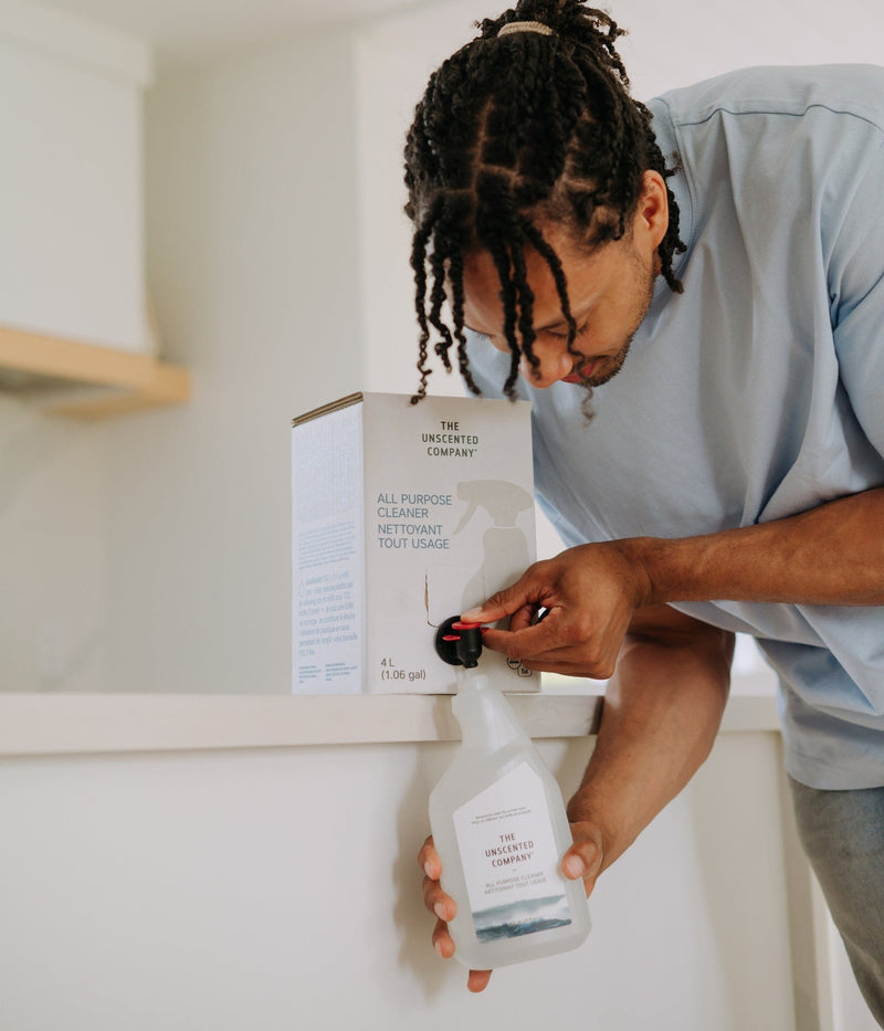 Man refilling an all-purpose cleaner spray bottle from a The Unscented Company 4L bulk refill station box in a bright, modern kitchen setting.