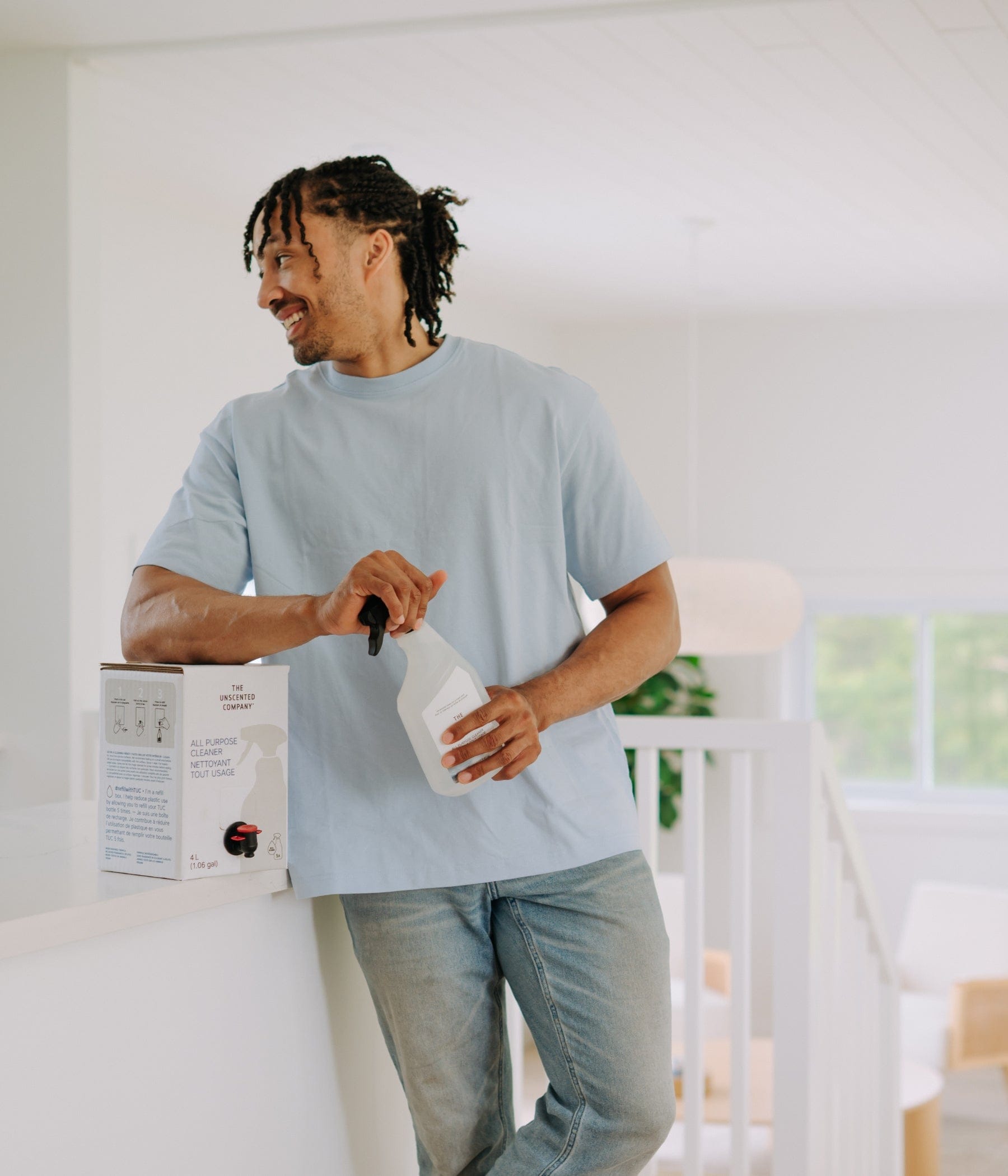 English
Man in a light blue t-shirt holding a spray bottle and a 4L bulk refill box of The Unscented Company all-purpose cleaner in a bright, modern interior.