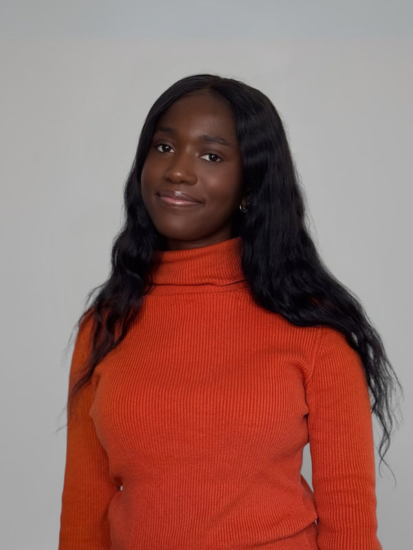 Portrait of Aissatou with long wavy black hair wearing a burnt orange turtleneck sweater, smiling softly against a neutral light gray background – professional studio headshot with natural lighting.