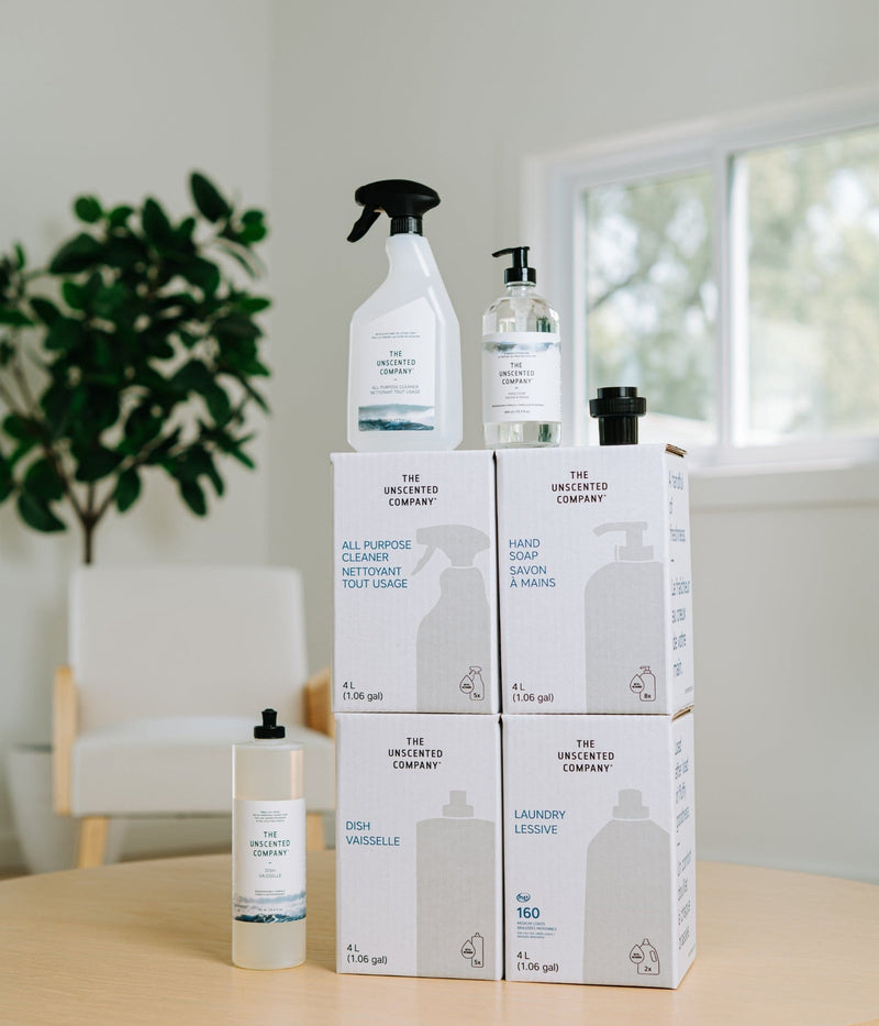 Photographie d'ambiance de la station de recharge à domicile The Unscented Company incluant des recharges en vrac de 4L de lessive et savons, disposées sur une table en bois dans un intérieur moderne et lumineux.