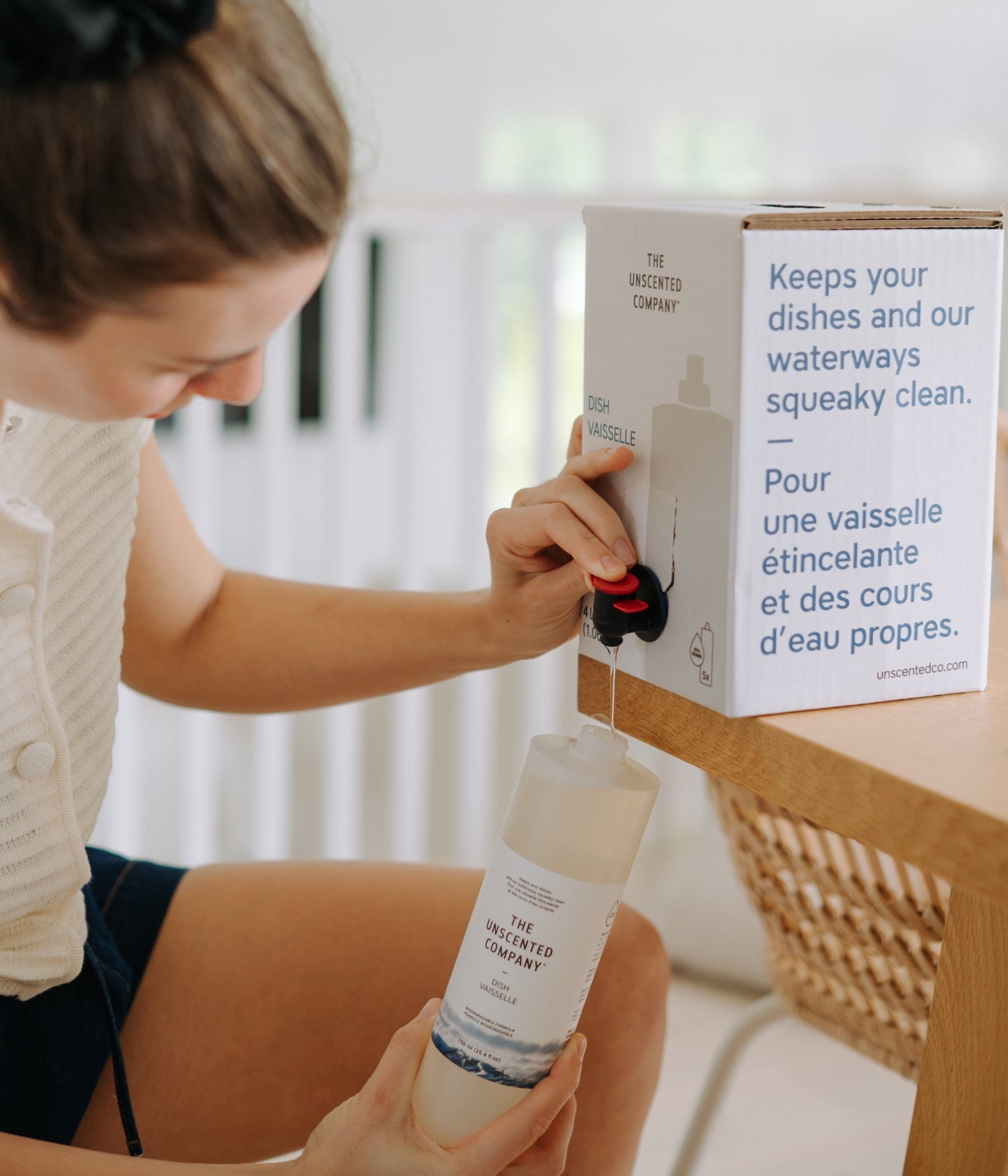 Woman refilling a dish soap bottle from a The Unscented Company 4L bulk refill station box on a wooden table in a bright home setting.
