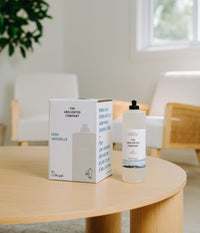 The Unscented Company 4 L dish soap bulk refill box and a reusable clear glass pump bottle displayed on a round wooden table in a bright, minimalist interior.