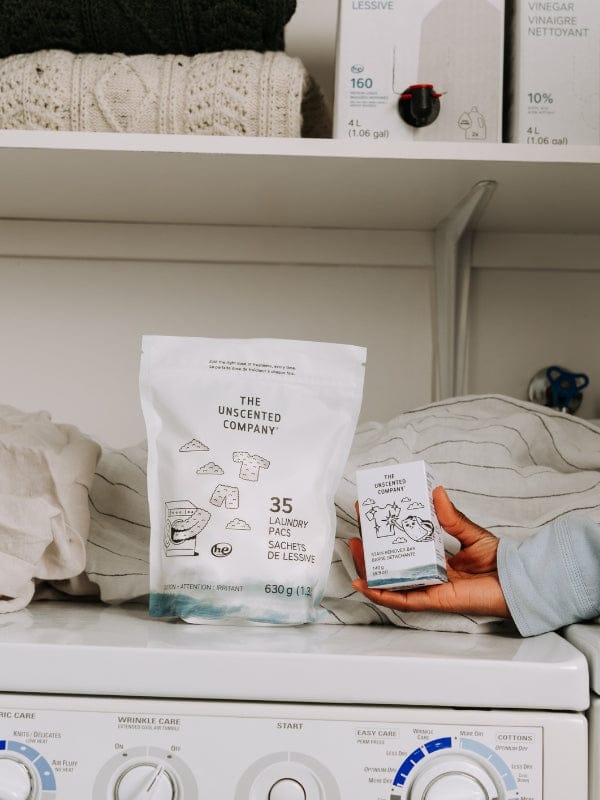 A bag of 35 laundry pacs (630 g) and a hand holding a stain remover bar box by The Unscented Company on a white washing machine. In the background, knit sweaters and bulk refill boxes (160 loads and 10% vinegar) are organized on a white shelf.