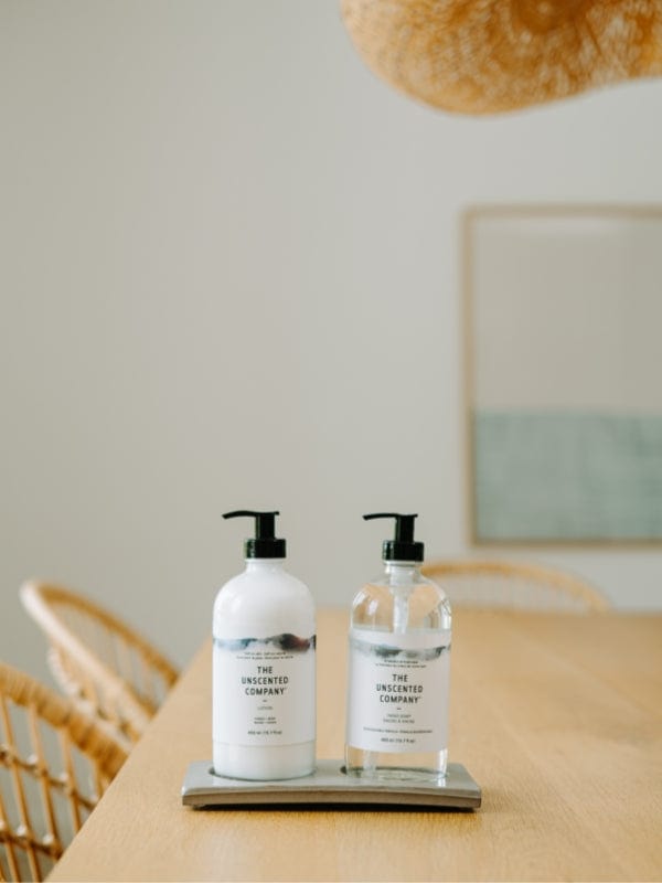 Two pump bottles of The Unscented Company products, one lotion and one hand soap, placed on a grey tray on a large light wood table, in a bright, minimal interior with rattan chairs and a minimalist framed artwork in the background.