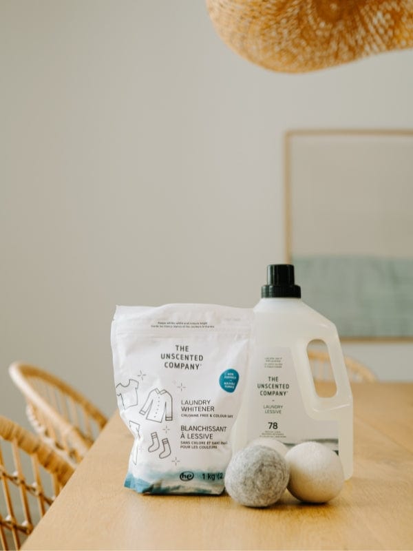 The Unscented Company laundry whitener and detergent placed on a wooden table alongside two wool dryer balls in a warm, minimalist interior.