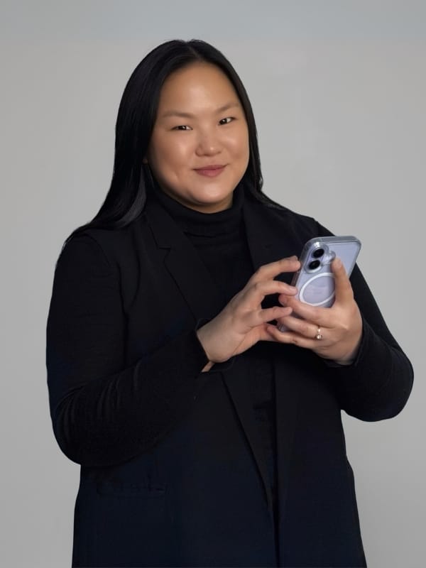 A woman with long black hair, wearing a black blazer over a black turtleneck, holds a light purple smartphone with a circular MagSafe case against a solid gray studio background.