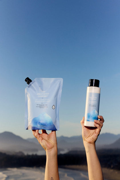 Two hands hold The Unscented Company hair conditioner refill pouch and matching bottle against a clear blue sky, with mountains in the distance.