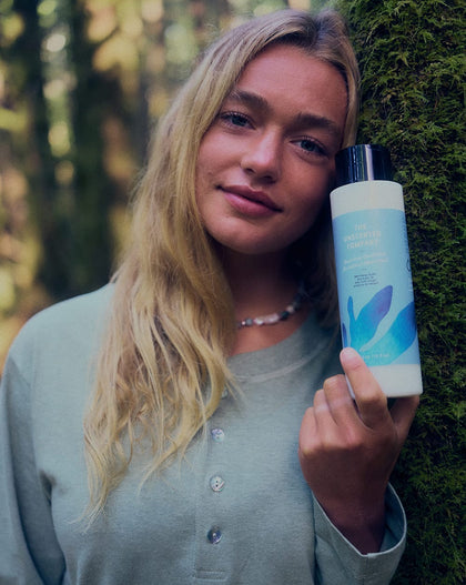A woman leaning against a moss-covered tree in a forest, holding The Unscented Company Nourishing Conditioner bottle close to her face.