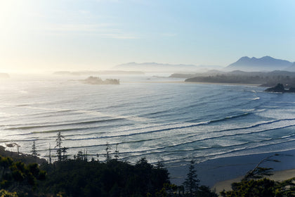 An aerial coastal landscape at sunrise showing rolling waves, a sandy beach, scattered forested islands, and layered mountain silhouettes fading into the mist.