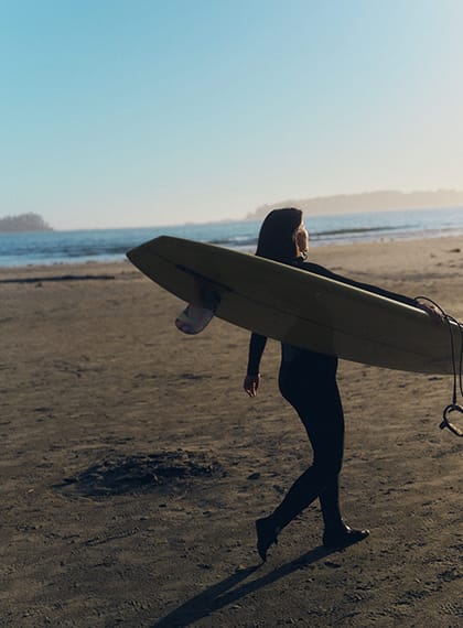 A surfer in a wetsuit walking along a sandy beach carrying a longboard, with the ocean and a clear sky in the background.
