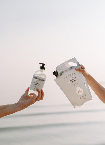 Two hands holding a hand soap bottle and refill pouch from The Unscented Company in front of the sea.