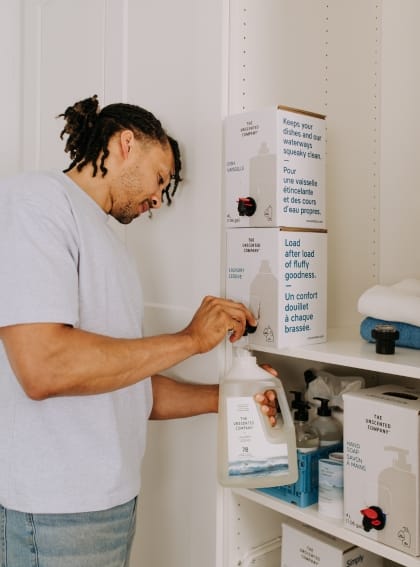 A person refilling a The Unscented Company laundry detergent bottle from a 4L refill box on a laundry room shelf, surrounded by other Unscented Company products including hand soap and dish soap.