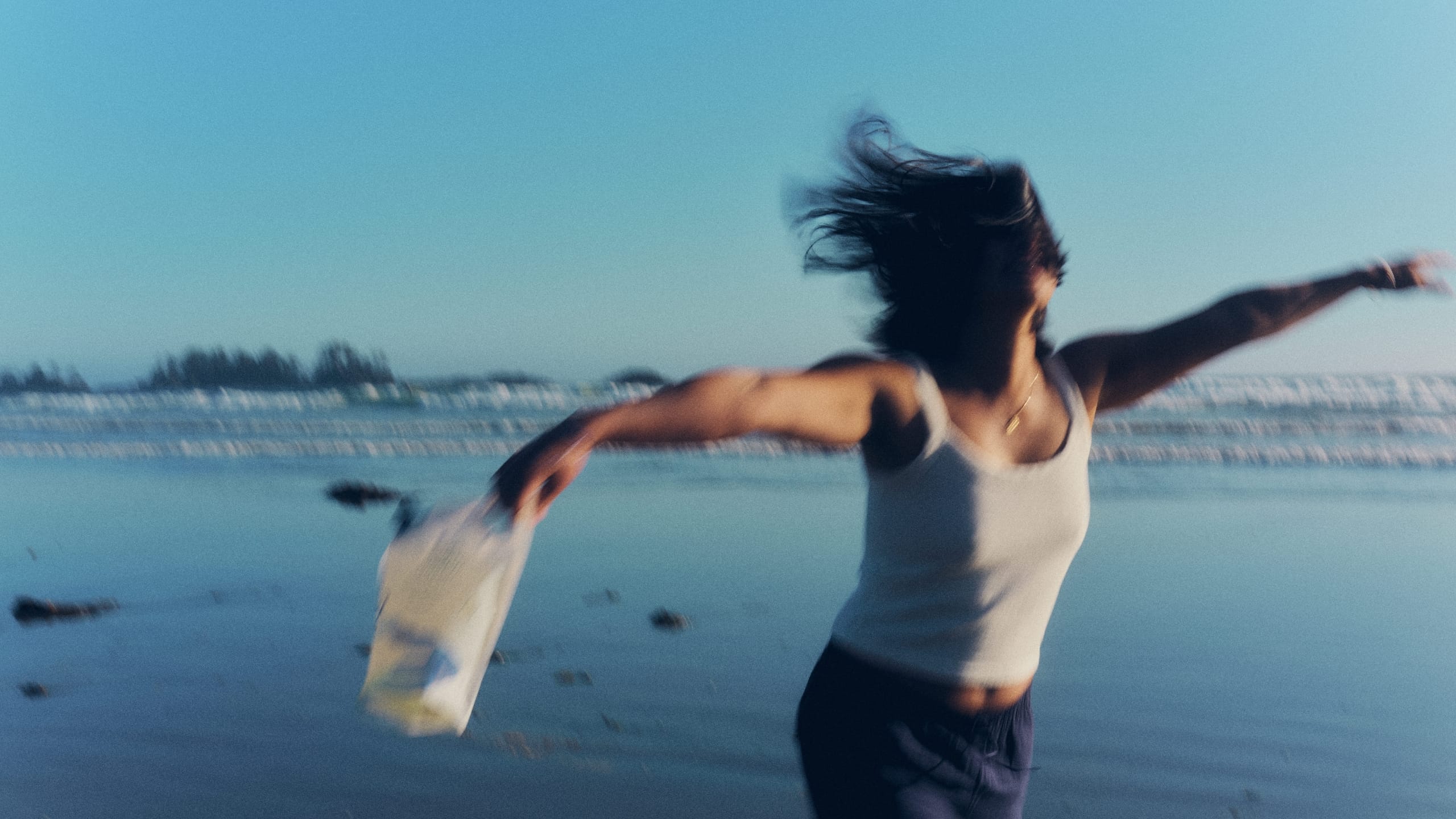 A motion-blurred image of a woman spinning on a beach at dusk, holding a The Unscented Company refill pouch with her hair flying in the wind.