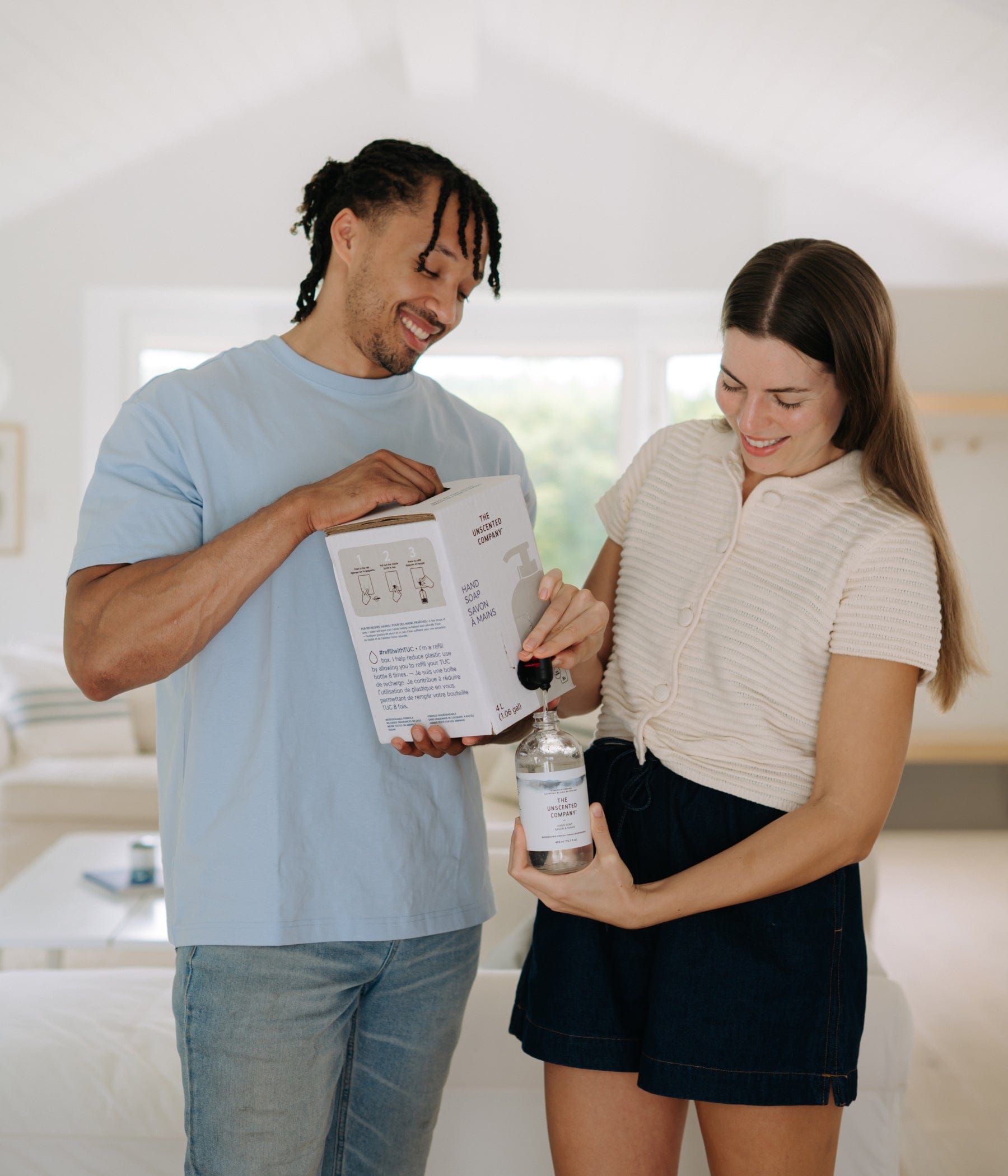 Couple unboxing The Unscented Company 4 L refill box indoors, pouring product into a reusable bottle in a bright, minimalist home setting.

