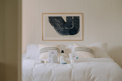 An assortment of The Unscented Company laundry products arranged on a minimalist white bed, with blue abstract wall art in the background.