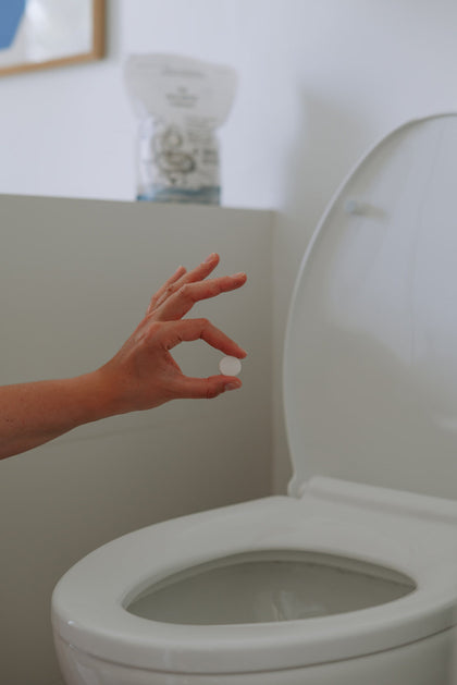 Close-up of a hand holding a toilet cleaning tablet above an open toilet bowl, ready to drop it in. A pouch of The Unscented Company toilet bowl cleaner is visible in the background.