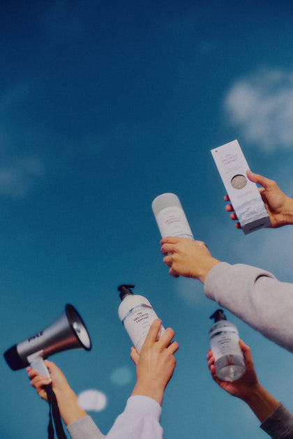 Several hands raise The Unscented Company products toward the blue sky, creating a vibrant and celebratory scene.