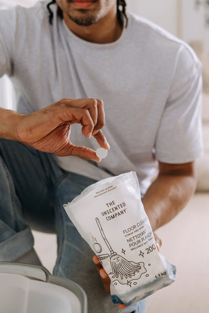 Close-up of a man’s hand holding a white cleaning tablet above a refill pouch of The Unscented Company floor cleaner. A bucket is partially visible in the corner.