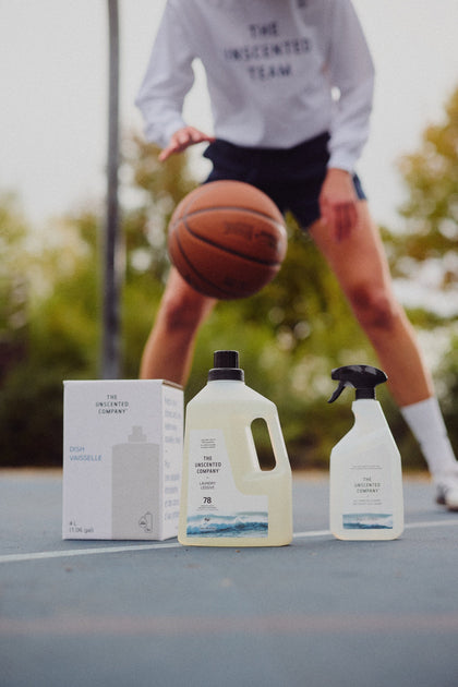 Three The Unscented Company products placed on an outdoor basketball court, with a person in motion bouncing a ball in the background.