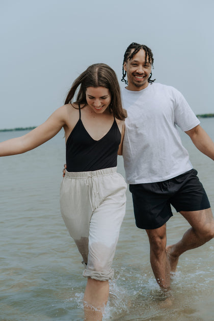 Un couple souriant marchant dans l'eau peu profonde d'un lac en été, illustrant un style de vie sain, minimaliste et écoresponsable.