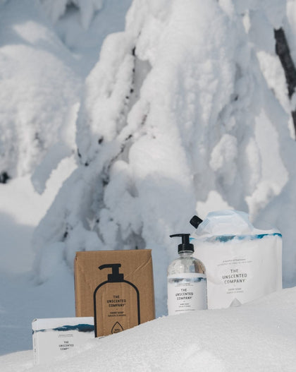 The Unscented Company products placed in the snow in front of heavily snow-covered trees, including a cardboard box, a hand soap bottle, a refill pouch, and a soap bar.