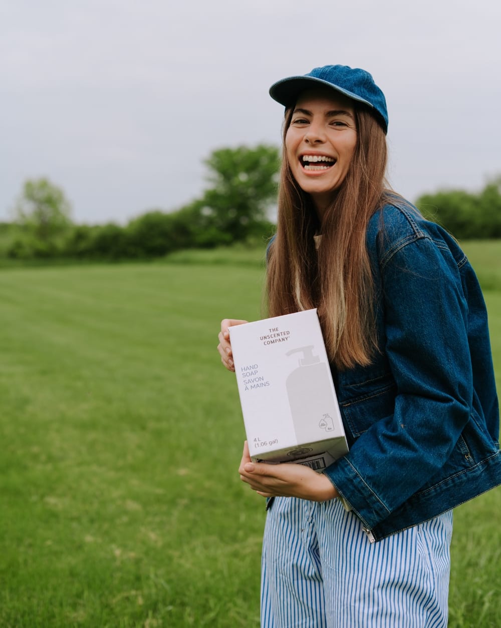 Smiling woman outdoors in a green field holding a 4L refill box of The Unscented Company Hand Soap, highlighting eco-friendly, fragrance-free personal care and sustainable refill packaging.
