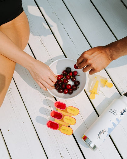 Vue de haut de mains partageant des cerises fraîches sur une terrasse en bois blanc, à côté d'une gourde réutilisable The Unscented Company et de moules à popsicles.