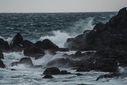Powerful waves crashing against black coastal rocks under a grey sky, capturing the raw energy of the ocean in motion.
