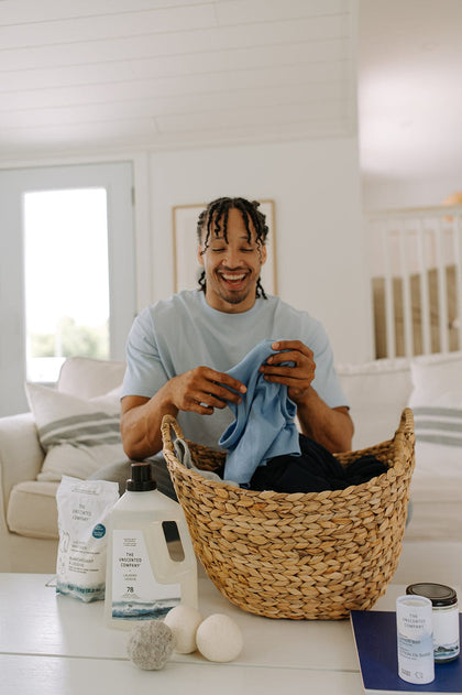 A smiling man looking at a clean garment over a wicker laundry basket, with detergent and dryer balls on a coffee table.