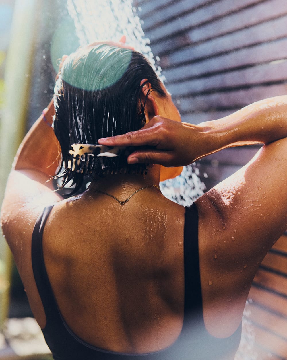A woman seen from behind washing her hair under an outdoor shower, using a hair clip to section her wet hair.