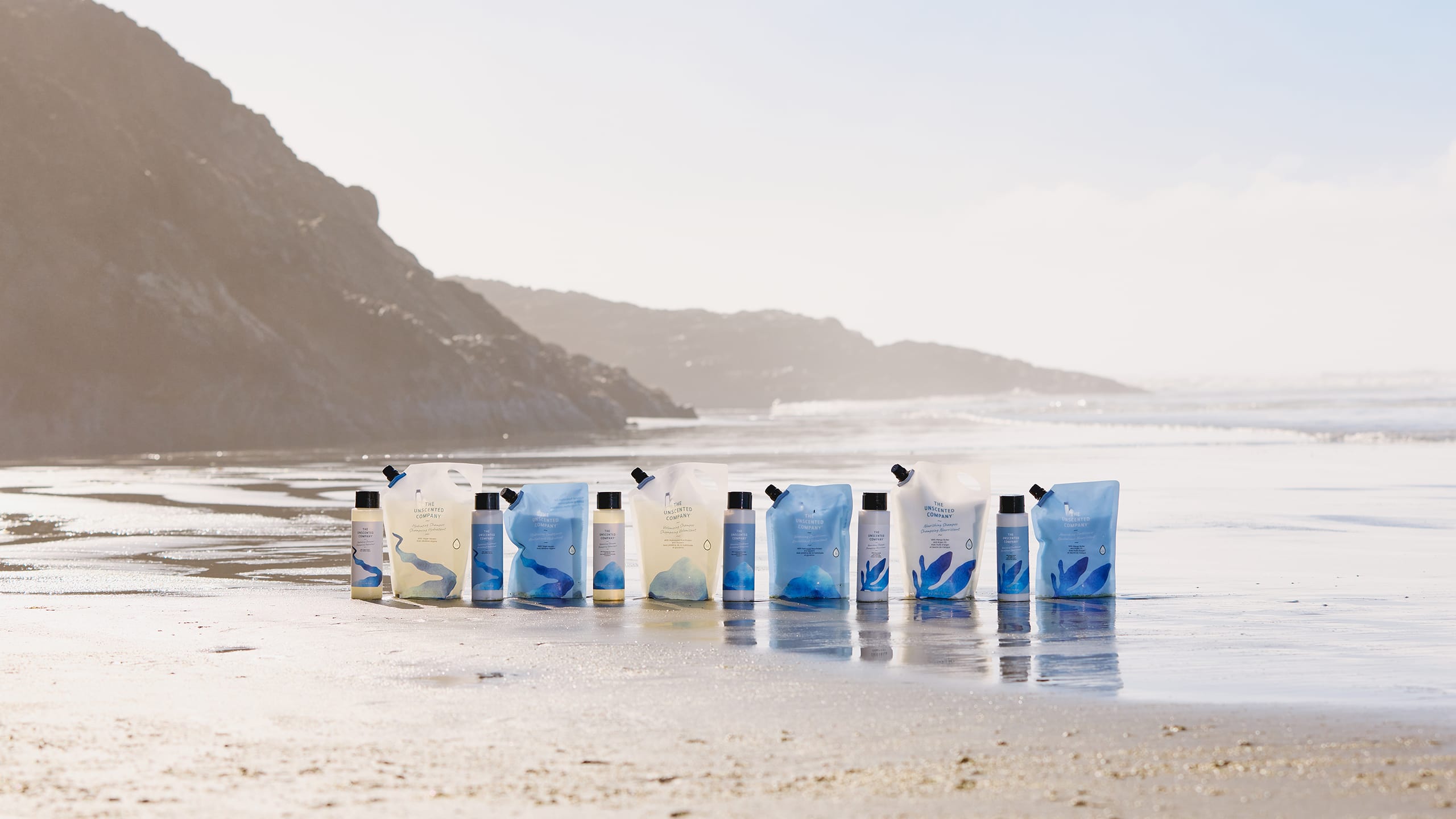 A collection of The Unscented Company shampoo, conditioner, moisturizing shampoo and hair care refill pouches with matching bottles arranged on wet sand at the beach, with ocean waves and rocky cliffs in the background.
