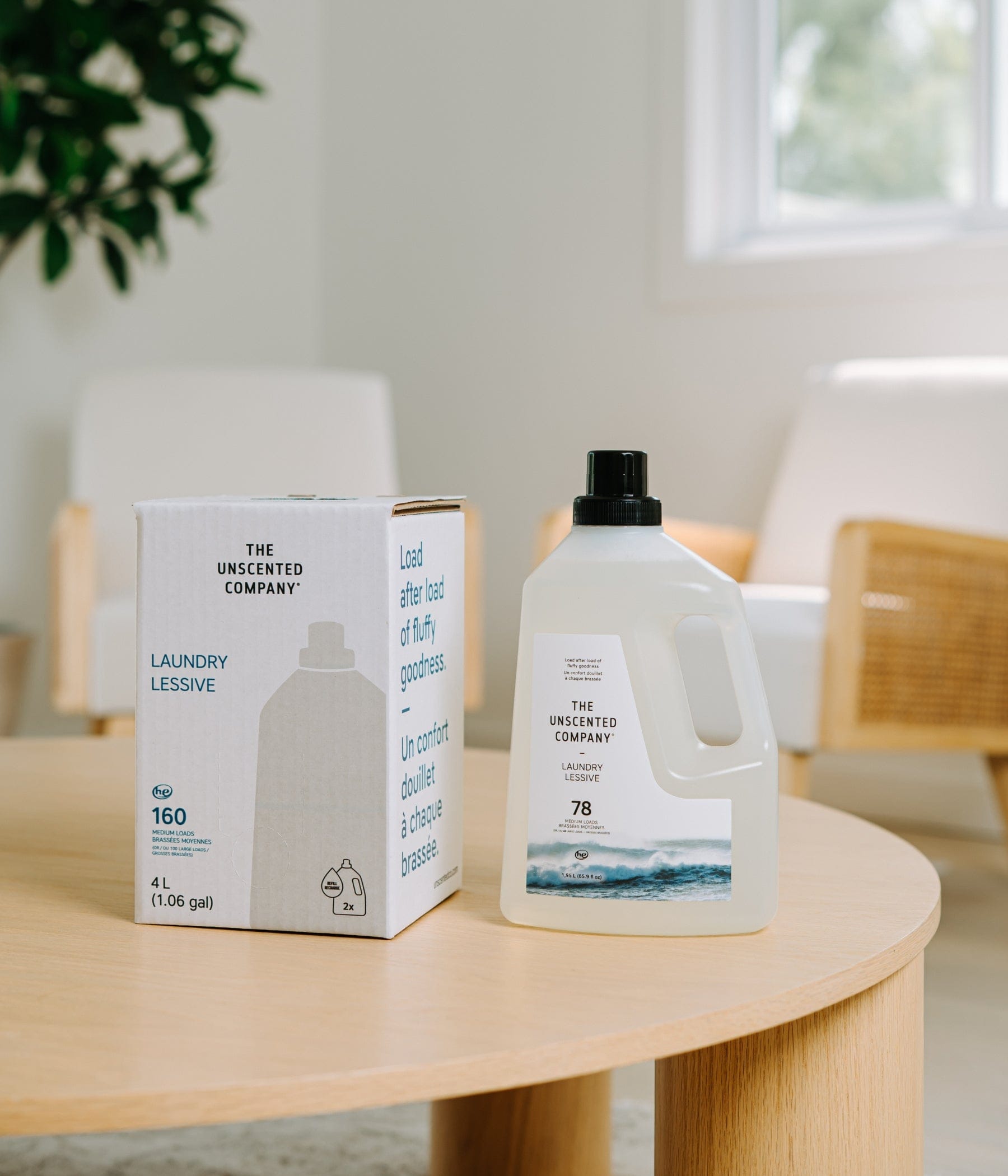 The Unscented Company 4L laundry detergent bulk refill box and a reusable white plastic detergent jug displayed on a round wooden table in a minimalist, bright interior.