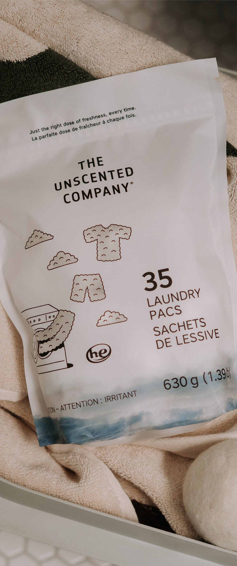 Vertical close-up of a 35-count bag of The Unscented Company laundry pacs (630 g) sitting in a laundry basket on a striped towel, with a wool dryer ball visible at the bottom.