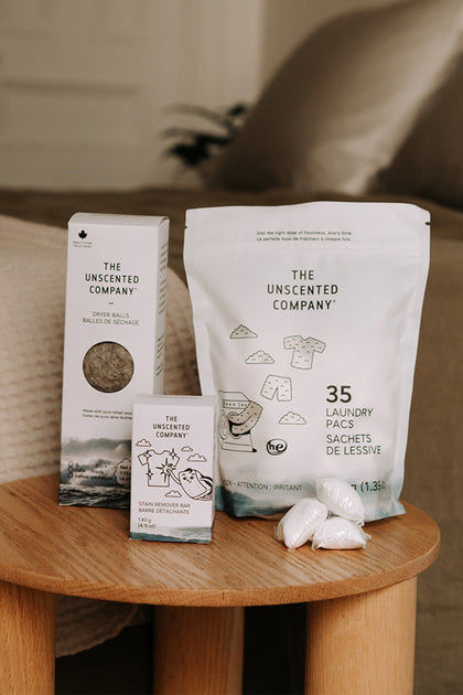 An assortment of products on a wooden table including a box of dryer balls, a stain remover bar, and a 35-count bag of laundry pacs with three loose pods next to it.