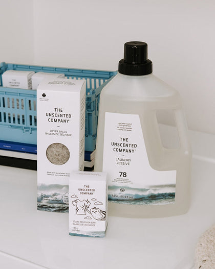 Close-up of a liquid laundry detergent jug (78 loads), a box of wool dryer balls, and a stain remover bar by The Unscented Company on a clean white surface.