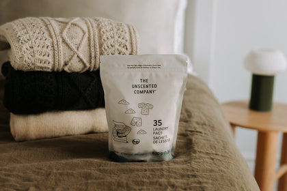 A white bag of 35 laundry pacs (630 g) by The Unscented Company sitting on an olive green linen bed next to a stack of three cozy knit sweaters in cream and forest green tones.