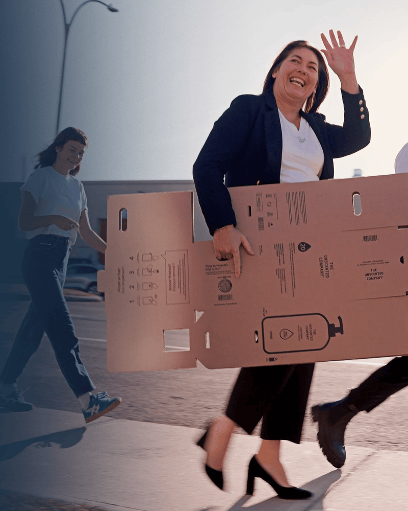 Anie Rouleau, founder of The Unscented Company, waving while walking with her team and carrying product packaging boxes, highlighting the brand’s growth and sustainable distribution.
