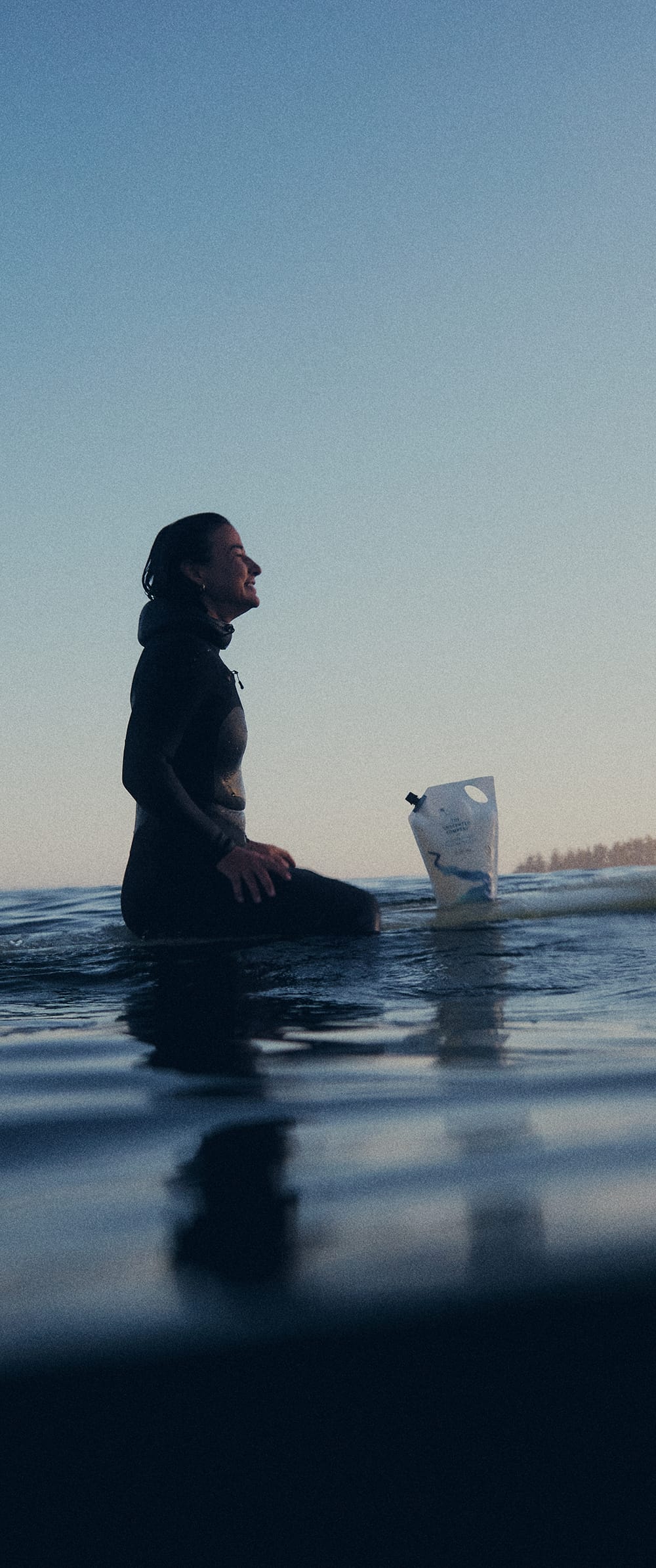 A woman in a black wetsuit sits on a surfboard in the ocean beside a The Unscented Company hair care refill pouch, smiling as gentle waves move around her.