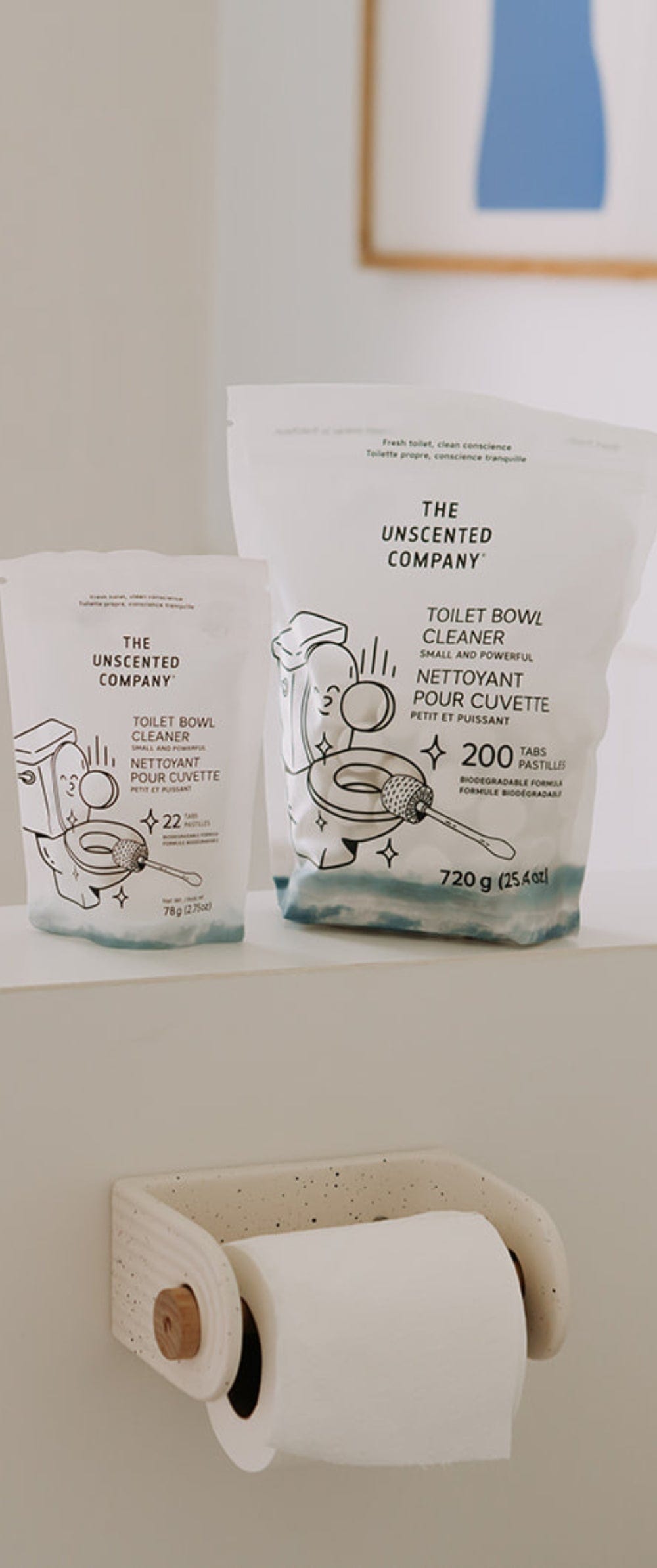 Two sizes of The Unscented Company toilet bowl cleaner bags are placed on the edge of a modern bathtub. A roll of toilet paper is mounted below, with a blue and white striped towel hanging in the background and a minimalist blue art print on the wall.