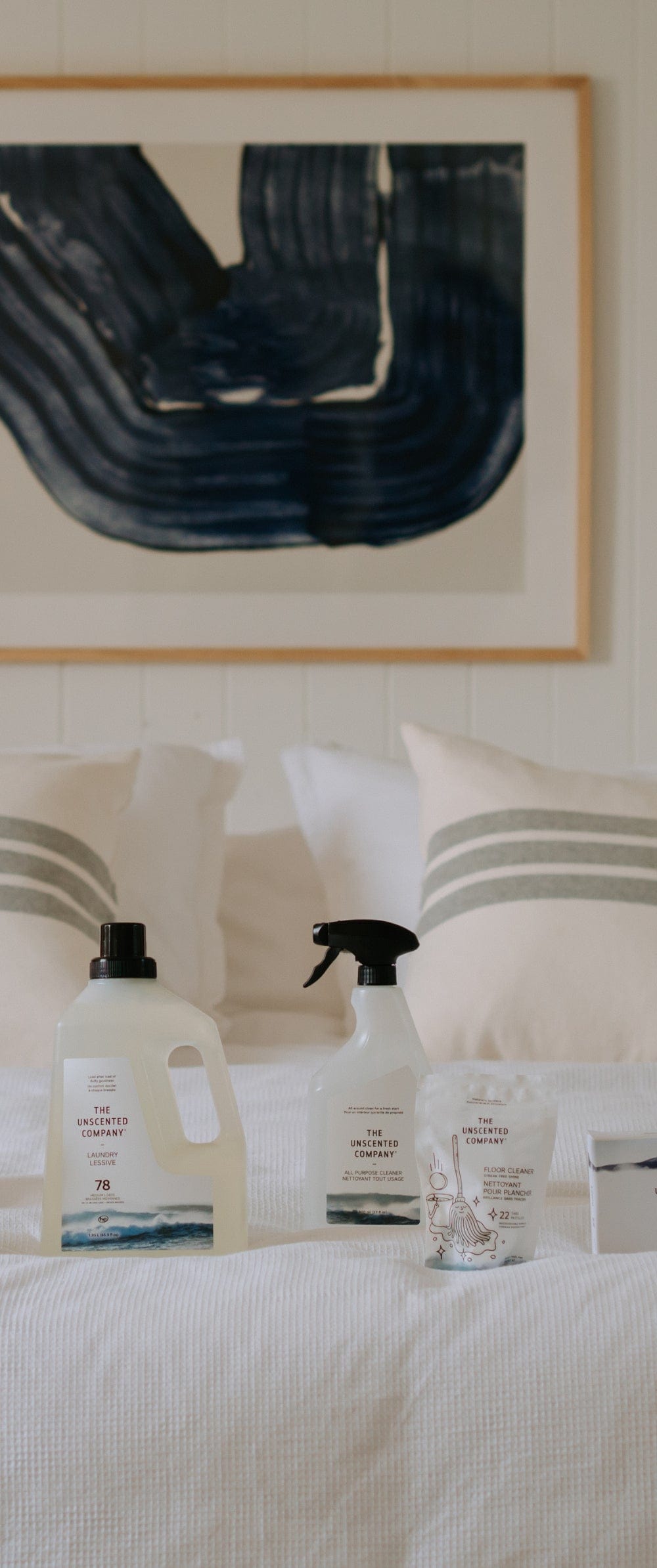 Close-up of The Unscented Company household products neatly arranged on a white bed in a bright, minimalist bedroom, with two striped pillows and a blue abstract artwork hanging on the wall in the background.