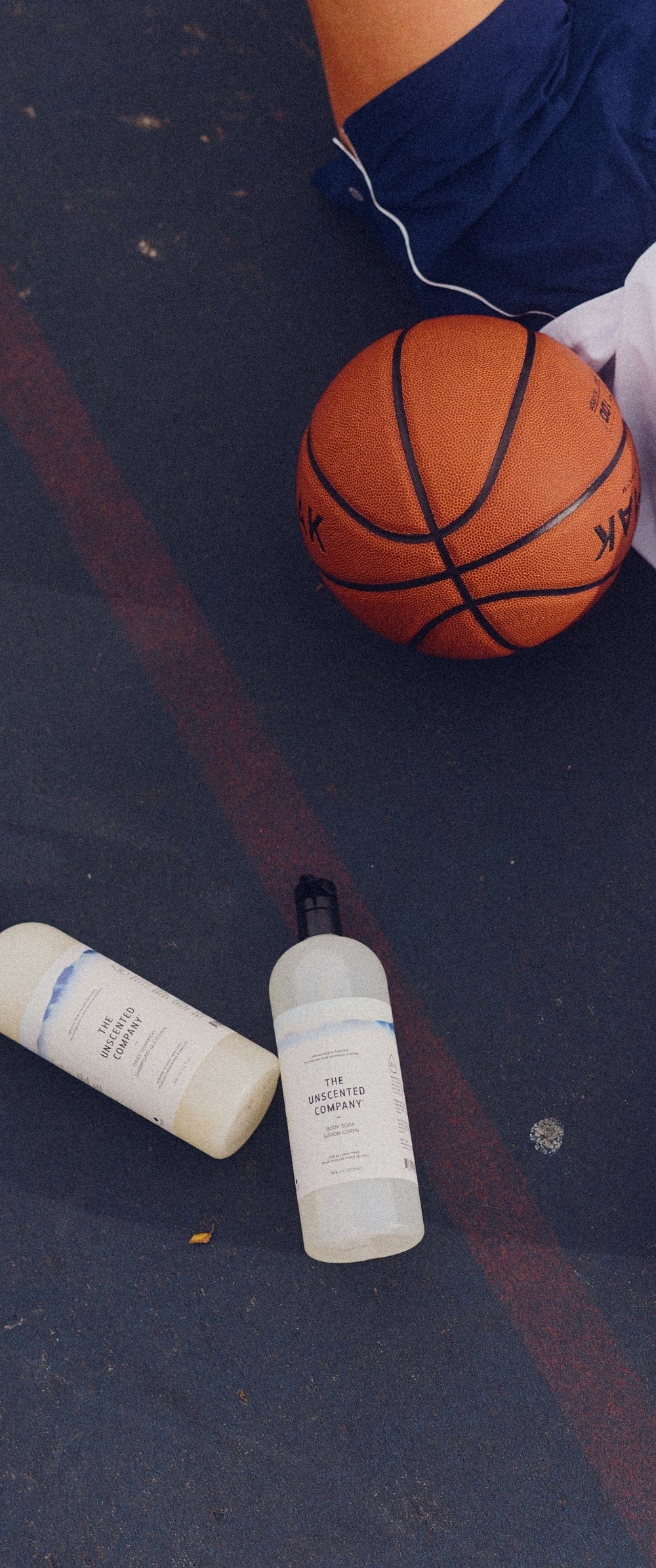 Two bottles of The Unscented Company body soap on a basketball court, next to a basketball and a seated person wearing blue shorts.